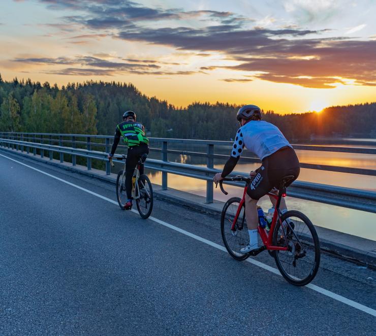Pyöräilijöitä takaa päin kuvattuna sillalla auringonlaskussa